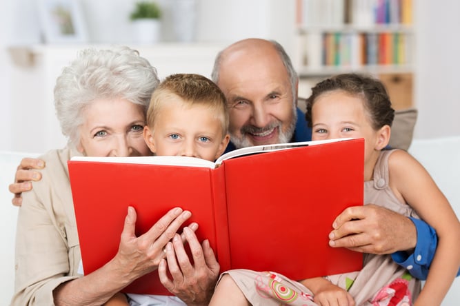 Cute little boy and girl with merry smiling eyes reading with their grandparents peering over the top of the book at the camera